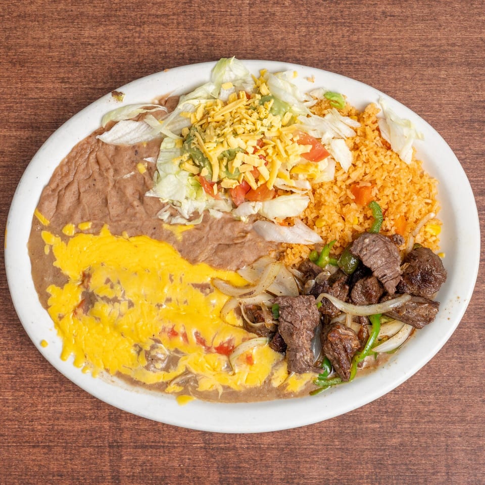 A white oval plate holds a Mexican dish with refried beans, rice, steak, and salad.
