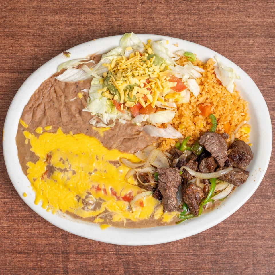 A white oval plate holds a Mexican dish with refried beans, rice, steak, and salad.
