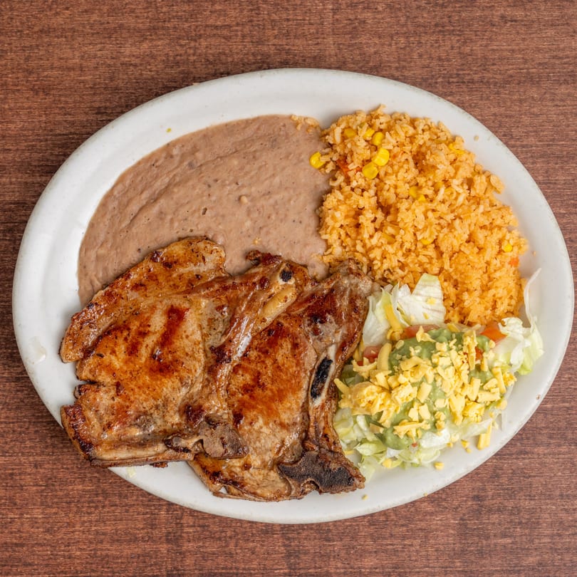 Two grilled pork chops with refried beans, rice, and a side salad with guacamole and cheese.