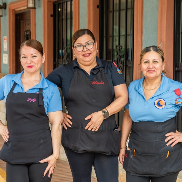 Three smiling women in blue shirts and black aprons stand outside a restaurant.