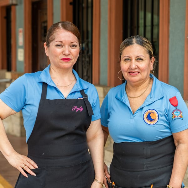 Two smiling women in blue polo shirts and black aprons stand outside a restaurant.