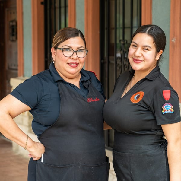 Two smiling women in black aprons and shirts stand side-by-side, looking at the camera.